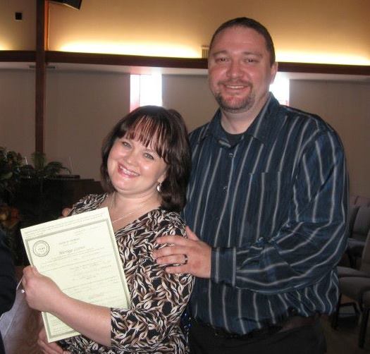 Heather and her late husband Steve stand close together in a softly lit church, smiling warmly at the camera. Heather is holding their marriage license, and Steve has one hand resting gently on her shoulder and the other on her arm. Both look happy and proud, captured on a meaningful, joyful day.