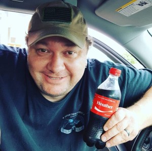 A man smiles warmly from the driver’s seat, holding a Coke bottle labeled ‘Heather.’ The image captures love, humor, and everyday joy from life before loss.