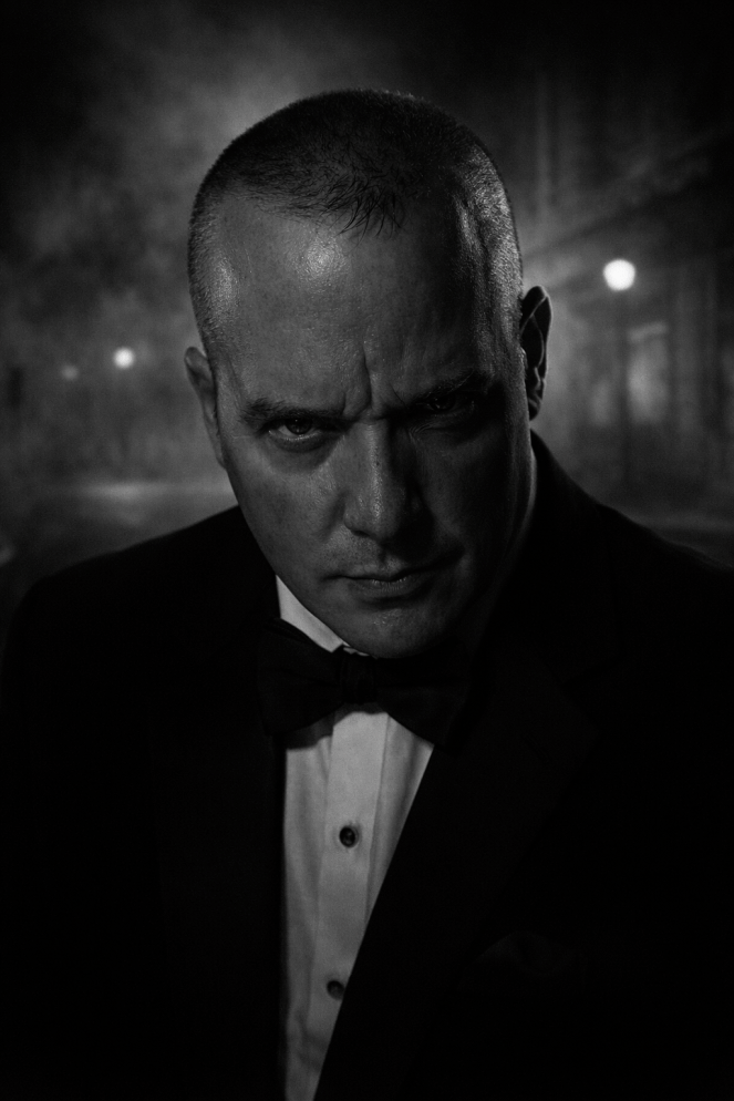 A dramatic black-and-white, film noir–style portrait of a middle-aged man in a tuxedo, staring intensely into the camera under low lighting. The image evokes classic crime drama and mystery, with a moody, ominous tone.