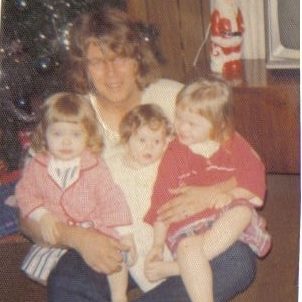Vintage 1970s Christmas photo of a father with long hair holding three young children near a Christmas tree in a family living room.
