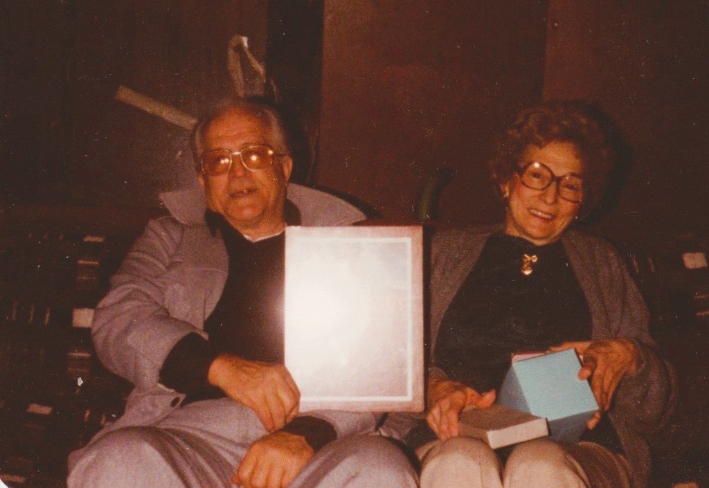An elderly couple seated indoors at Christmas, smiling gently while holding wrapped gifts. The photo has a warm, slightly faded look typical of the mid-1980s.