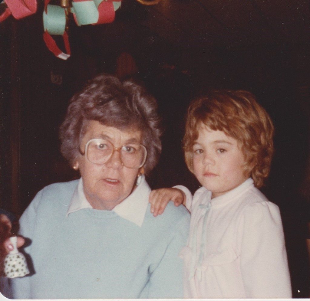 An elderly woman with short gray hair and glasses sits beside a young girl with light brown hair, the child’s hand resting on the woman’s shoulder. They are indoors at Christmas, with paper chain decorations hanging above them.