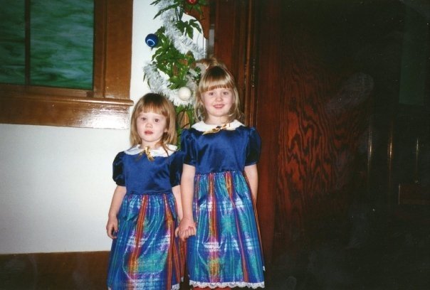 Two young girls stand side by side in matching blue Christmas dresses with rainbow-patterned skirts, holding hands in front of a decorated Christmas tree at a church program in the mid-1990s.