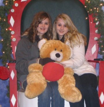 Two sisters sit close together at a Christmas light display, smiling softly while holding a large teddy bear, surrounded by glowing holiday lights.