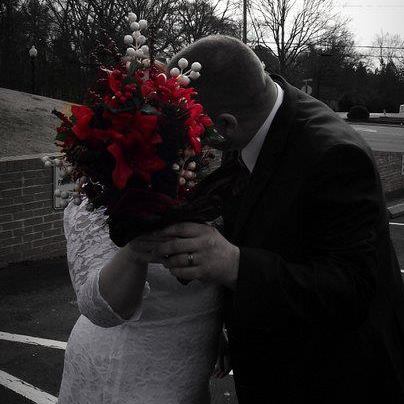 A black-and-white photo of a bride and groom kissing outdoors after their winter wedding ceremony, with the bride holding a bouquet of red flowers.
