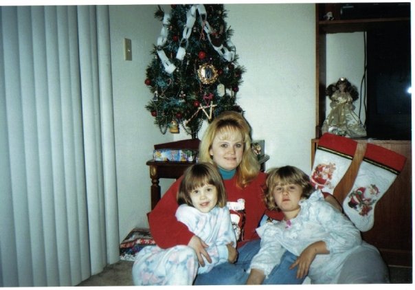 A young mother sits on the floor in front of a decorated Christmas tree, holding her two small daughters on her lap. The children are wearing cozy pajamas, surrounded by wrapped gifts and stockings, capturing a quiet family moment from Christmas 1997.