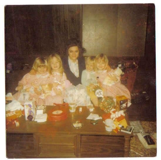 A faded 1970s Christmas photo showing a mother seated behind a coffee table with her young children gathered closely around her, surrounded by opened gifts and wrapping paper, capturing a warm, slightly chaotic holiday morning.