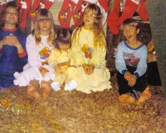 Vintage 1970s Christmas photo of five siblings sitting barefoot on a shag carpet in front of a fireplace decorated with stockings, wearing cozy pajamas and looking tired, excited, and perfectly imperfect.